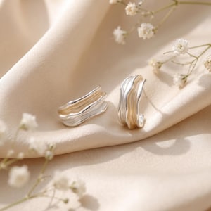 May include: A pair of silver-toned hoop earrings featuring a distinct wavy, rippled texture. The earrings have a polished finish with curved, organic edges that create a layered appearance, reflecting light across their metallic surfaces.