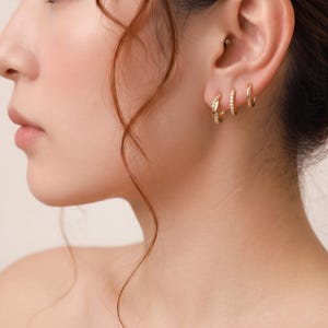 Könnte beinhalten: Drei goldene Creolen, die an einem Ohrläppchen getragen werden. Die erste Creole hat ein gedrehtes Design, die zweite ist ein dünner Ring, der mit kleinen klaren Steinen besetzt ist, und die dritte ist eine schlichte, glatte goldene Creole.