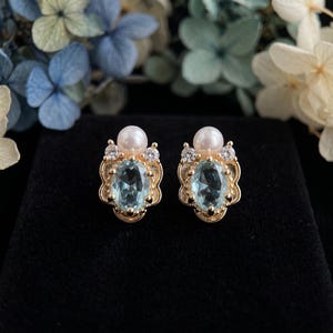 May include: A pair of gold stud earrings featuring a central oval-cut light blue gemstone, a round white pearl positioned above the stone, and two small clear round-cut accent stones on either side of the pearl, set in a decorative gold frame.