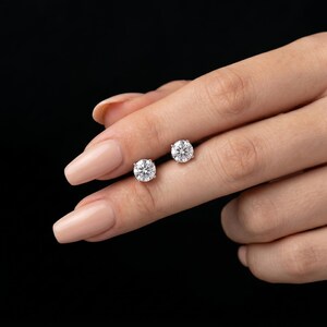 May include: A pair of sparkling, round-cut diamond stud earrings. The earrings are held on a person's finger against a black background. The diamonds are clear and reflect light, showcasing their brilliance.