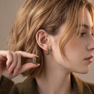 Puede incluir: Un par de pendientes de aro dorados con una hilera de pequeñas piedras de color verde oscuro incrustadas en la parte frontal. Un pendiente se lleva en el lóbulo y el otro se sostiene entre los dedos para mostrar su textura y color.