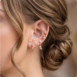 May include: A set of five gold-toned earrings worn in a single ear. The collection includes a small planet charm, a tiny star, a heart-shaped stud, a small bear charm with a pink stone, and two small studs featuring light pink circular stones.