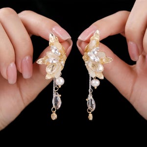 Può includere: Un paio di orecchini dorati con una base testurizzata a forma di foglia, decorati con cristalli trasparenti taglio marquise e pietre opache color crema. Ogni orecchino include una perla bianca pendente, una goccia di cristallo sfaccettato e un piccolo ciondolo dorato.