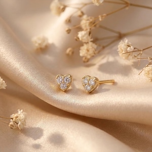 May include: A pair of gold-colored heart-shaped stud earrings with small, clear gemstones. The earrings are displayed on a satin surface with dried flowers in the background. The earrings are a romantic jewelry item.