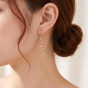 Puede incluir: Un pendiente de hilo de oro con un delicado diseño en espiral adornado con pequeñas piedras claras y brillantes. El pendiente consiste en una fina cadena de oro pulido que se pasa por el lóbulo de la oreja y termina en una espiral metálica.