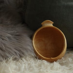 May include: A small, round, light-coloured wooden bowl with a smooth, polished interior and a distinct, carved handle protrusion at the top rim. The bowl features a natural wood grain pattern and a warm, golden-brown hue throughout its surface.