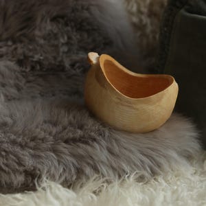 May include: A small, light-coloured wooden bowl with a smooth, rounded exterior and a curved, asymmetrical rim. The bowl features a natural wood grain pattern and a light tan finish, showcasing a simple, functional design with a small handle protrusion.