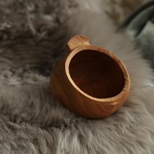 May include: A small, round, light brown wooden cup with a smooth, polished finish and a single, integrated handle tab at the top rim. The cup features a deep, hollow interior and a rounded base, showcasing the natural grain patterns of the wood.