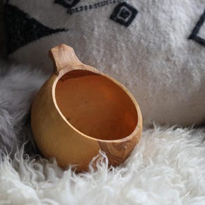 May include: A light brown wooden kuksa cup with a smooth, rounded bowl and a single protruding handle. The cup features visible natural wood grain patterns and a polished finish, showcasing the organic texture and warm tones of the carved wood.