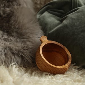 May include: A small, round wooden cup with a single handle, featuring a natural light brown wood grain finish. The cup has a smooth, polished interior and a sturdy, thick-walled construction, showcasing the organic texture of the carved wood.