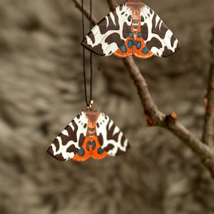 May include: Two moth-shaped ornaments hanging from thin black cords. Each ornament features a detailed pattern of dark brown and white wings with vibrant orange and blue accents near the center, suspended from a thin, bare wooden branch.