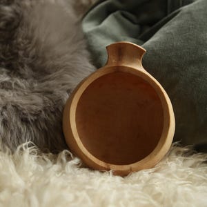May include: A round, light-coloured wooden cup with a smooth, polished finish and a single integrated handle extending from the top rim. The cup features a deep, circular basin and a natural wood grain pattern visible across its curved exterior surface.