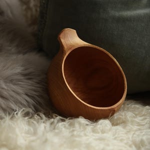 May include: A small, round, light brown wooden cup with a single handle protruding from the top rim. The cup features a smooth, polished interior and a natural wood grain texture, showcasing a simple, functional design crafted from solid wood.