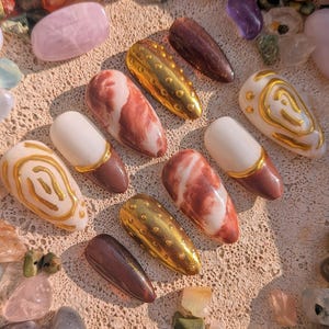 Könnte beinhalten: Eine Sammlung künstlicher Fingernägel in verschiedenen Designs und Farben. Einige Nägel sind weiß mit braunen Spitzen, andere marmoriert rot und weiß, und einige sind einfarbig braun oder gold. Die Nägel sind von bunten Edelsteinen umgeben.