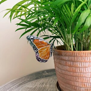 Può includere: Una farfalla decorativa con ali arancioni e nere è attaccata a una pianta verde in un vaso di terracotta. La farfalla ha un design dettagliato con contorni e accenti neri. La pianta ha foglie lunghe e verdi.