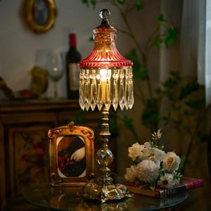 May include: An ornate table lamp featuring a red and amber glass shade, crystal droplets, and a gold-toned metal base. The lamp is displayed on a glass table, alongside a framed picture and a bouquet of white flowers. The lamp's height is approximately 30 cm.