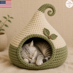 May include: A cream and sage green crocheted cat bed shaped like a pointed hood with a curled top. The entrance is trimmed in sage green with two crocheted leaves and a small brown button attached to the side. A grey tabby cat is sleeping inside.