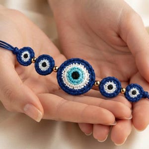 May include: A bracelet featuring five circular crocheted evil eye motifs in shades of dark blue, white, and light blue, connected by small metallic gold beads on a dark blue cord with adjustable ends finished with gold beads.