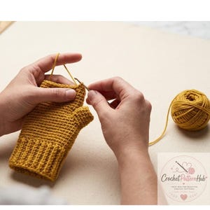 Puede incluir: Un guante sin dedos de ganchillo color amarillo mostaza con puño acanalado y abertura para el pulgar, trabajado con un ganchillo de metal y un ovillo de lana a juego. La imagen incluye un logotipo con el texto Crochet Pattern Hub.