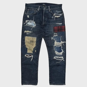 Op de afbeelding: Donkerblauwe denim jeans met distressed details, waaronder scheuren en patches. Voorzien van een rood-zwarte geruite patch en andere stoffen patches. De jeans hebben een knoopsluiting en een klassiek vijf-zakken ontwerp.