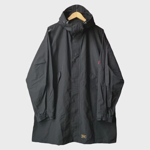 Peut inclure: Veste anorak noire à capuche avec fermeture éclair intégrale. La veste a une coupe ample, des manches longues et un petit accent rouge sur la manche droite. La veste est suspendue à un cintre en bois.