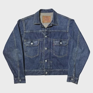 Op de afbeelding: Een vintage blauwe spijkerjas met twee borstzakken en knoopsluitingen. De jas heeft een klassieke kraag en een klein rood label op een zak. De denim heeft een versleten uitstraling.