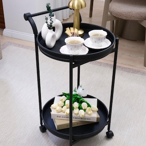 Peut inclure: Un chariot de service noir à deux niveaux avec une lampe dorée, des vases blancs, des tasses à thé et des livres. Le chariot a une poignée et des roues. L'étagère inférieure contient des livres et des tulipes blanches. L'étagère supérieure contient des tasses à thé et une lampe dorée.