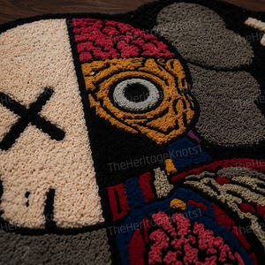 May include: A black, beige, red, and blue tufted rug with a skull and anatomical design. The skull has a black 'X' for eyes. The other side of the rug features an eye and brain design. The rug is made of a soft, textured material.