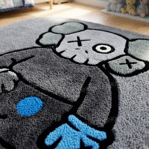 K&ouml;nnte beinhalten: Grauer Pl&uuml;schteppich mit einem Cartoon-Charakter-Design. Der Teppich hat einen grauen Hintergrund mit einer gro&szlig;en Figur in Grau-, Schwarz- und Blaut&ouml;nen. Die Figur hat kreuzf&ouml;rmige Augen und einen runden blauen Punkt.