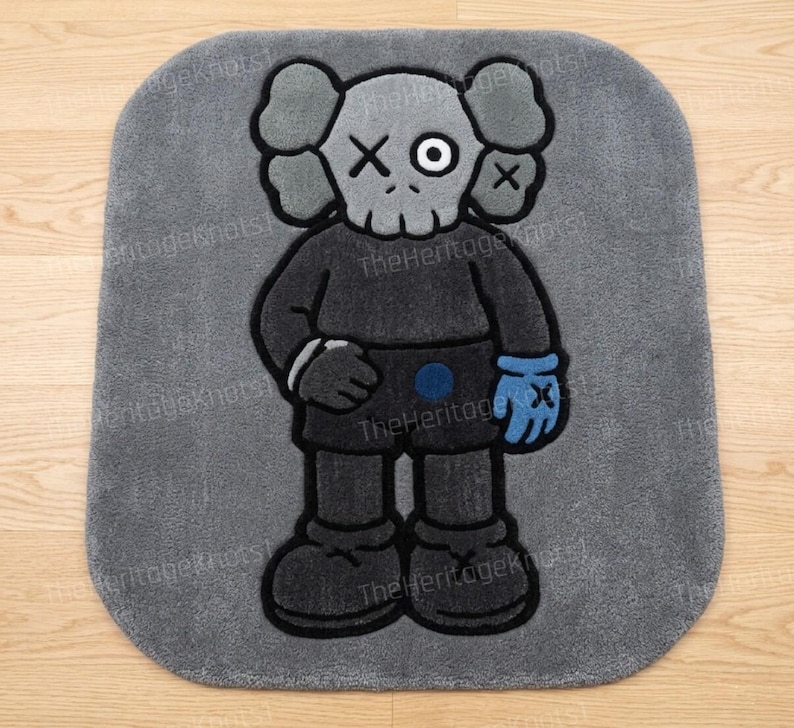 K&ouml;nnte beinhalten: Grauer rechteckiger Teppich mit einer Cartoonfigur mit sch&auml;del&auml;hnlichem Gesicht, X-f&ouml;rmigen Augen und einem blauen Handschuh. Die Figur tr&auml;gt ein dunkelgraues Outfit mit schwarzen Umrissen. Der Teppich hat abgerundete Ecken.