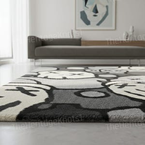 May include: A gray and white patterned area rug with a repeating cartoon-like face design. The rug has a plush texture and is placed on a light gray floor. The background includes a gray sofa and abstract artwork.