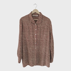 Op de afbeelding: Een shirt met lange mouwen met een geometrisch patroon van kleine, donkerrode vierkanten op een beige achtergrond. Het shirt hangt aan een houten hanger tegen een witte achtergrond. De merknaam "FRANCESCA" is zichtbaar op de kraag.