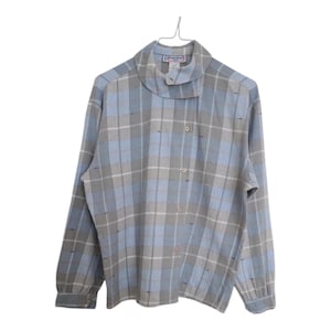 Può includere: Una camicia a quadri a maniche lunghe nei toni del blu, grigio e bianco, appesa a una gruccia di metallo. La camicia presenta un esclusivo collo a cascata e una chiusura con bottoni. Il tessuto ha un aspetto morbido e strutturato.