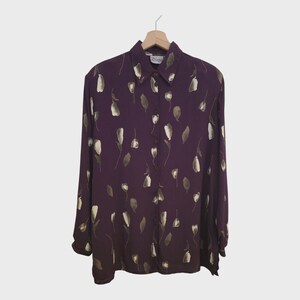 Può includere: Una camicia abbottonata a maniche lunghe in una tonalità viola intenso, ornata da un motivo floreale nei toni del bianco e del talpa. La camicia è appesa a una gruccia di legno, mostrando il suo colletto classico e il davanti abbottonato. L'etichetta recita "Dizzy".