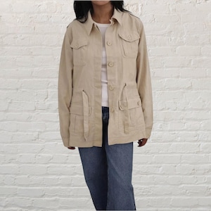 Può includere: Giacca utility beige con più tasche, chiusure con bottoni e collo. La giacca ha maniche lunghe e vita con coulisse. La modella indossa jeans blu e una maglietta bianca sotto.