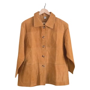 Könnte beinhalten: Eine hellbraune Wildlederjacke mit Kragen und langen Ärmeln. Die Jacke hat vier braune Knöpfe auf der Vorderseite und zwei Taschen. Sie hängt an einem Holzbügel vor weißem Hintergrund.