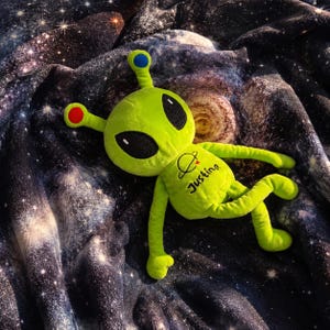 Puede incluir: Un juguete de peluche alienígena de color verde lima brillante con grandes ojos ovalados negros, dos antenas con puntas circulares azul y roja, y el nombre Justina bordado en letra cursiva negra en el pecho junto a un pequeño gráfico de un planeta.