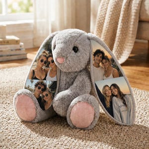 Könnte beinhalten: Ein graues Plüschkaninchen mit rosa Innenohren und Pfotenpolstern. Die Ohren des Kaninchens zeigen mehrere Fotos von Personen. Das Kaninchen sitzt auf einem beigefarbenen Teppich.