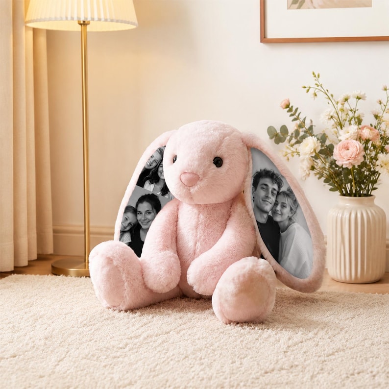 K&ouml;nnte beinhalten: Ein rosa Pl&uuml;schhase mit langen Ohren, der auf einem cremefarbenen Teppich sitzt. Die Innenseite jedes Ohres zeigt ein Schwarz-Wei&szlig;-Foto. Der Hase hat eine rosa Nase und eine weiche, flauschige Textur.