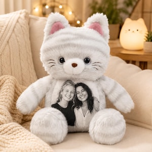 Può includere: Un peluche a forma di gatto con pelliccia a righe grigio chiaro e bianco, orecchie rosa all'interno e grandi occhi scuri. Il centro del busto del gatto presenta una fotografia in bianco e nero di due persone sorridenti stampata sul tessuto.