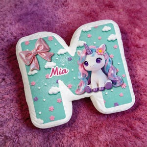 Op de afbeelding: Een decoratieve letter 'M' met een turquoise achtergrond, versierd met een roze strik, wolken en bloemen. Een eenhoorn met roze en blauw haar zit rechts. De naam 'Mia' staat op de letter.