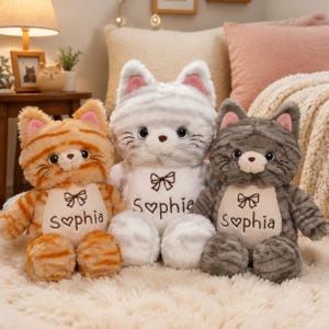 Peut inclure: Trois peluches en forme de chat aux motifs tigrés orange, gris clair et gris foncé. Chaque chat présente un empiècement blanc sur la poitrine brodé d'un nœud marron et du prénom Sophia, avec un cœur remplaçant la lettre o.