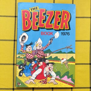 May include: A hardback book titled The Beezer Book 1976 with a blue cover featuring a colourful cartoon illustration of three characters in a field, one firing a toy gun and another holding a fishing rod with an ice cream cone dangling from the line.