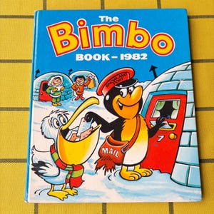 May include: A hardback book titled The Bimbo Book-1982 with a blue cover featuring a cartoon penguin wearing a Snowland Post hat and a mail bag, a pelican with a scarf holding letters, and igloos in a snowy landscape with two small figures.