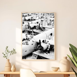 Peut inclure: Une impression encadrée en noir et blanc d'une photographie d'époque représentant l'assemblage d'avions. L'impression est accrochée au-dessus d'un bureau en bois avec une chaise blanche. Une grande plante en pot et un panier décoratif complètent le décor.