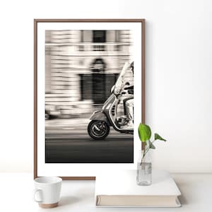 May include: A framed black and white photograph depicting a scooter in motion on a city street. The image features a blurred architectural background, a rider on the scooter, and a clear view of the front wheel and body of the vehicle.