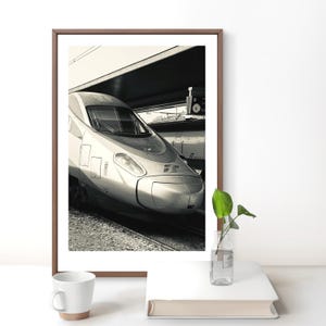 Peut inclure: Une photographie encadrée en noir et blanc montrant le nez d'un train à grande vitesse à quai. Le train présente un design aérodynamique épuré avec un phare proéminent et le logo des Ferrovie dello Stato Italiane sur le panneau avant.
