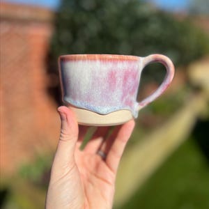 Könnte beinhalten: Eine handgefertigte Keramiktasse mit einer einzigartigen Glasur. Die Tasse hat einen hellbeigen Boden, der in einen weichen rosa-weißen Körper und einen rot-orangen Rand übergeht. Der Griff ist gebogen. Die Tasse wird in einer Hand gehalten.
