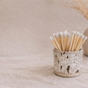 Op de afbeelding: Een gespikkelde keramische luciferhouder bevat lucifers met witte uiteinden. Op de achtergrond staat een kleine, getextureerde vaas met gedroogd gras. De scène speelt zich af op een neutraal getinte ondergrond.