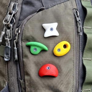 Peut inclure: Un sac à dos vert olive foncé et noir avec quatre petites prises d'escalade fixées sur la poche avant. Les prises sont en forme d'étoile blanche à quatre branches, de rebord vert incurvé, d'ovale jaune percé et de demi-cercle rouge.